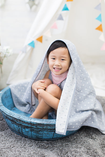 toddler girl smiles wearing GOTS certified organic cotton pink bandana bib and gray blanket on her head